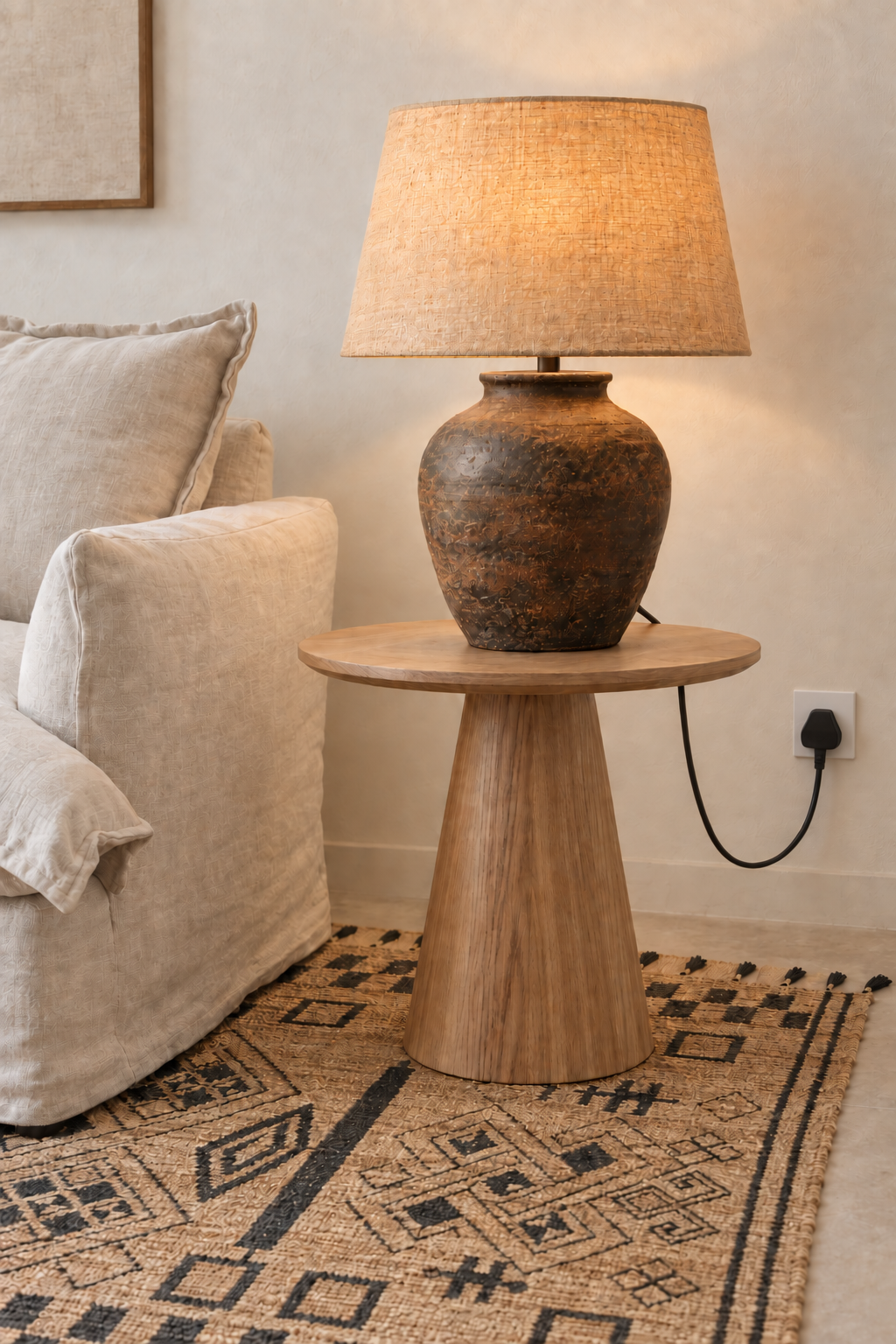 A rustic table lamp with a textured brown ceramic vase base and a beige woven drum shade, sitting on a round wooden side table in a living room next to a light beige sofa.