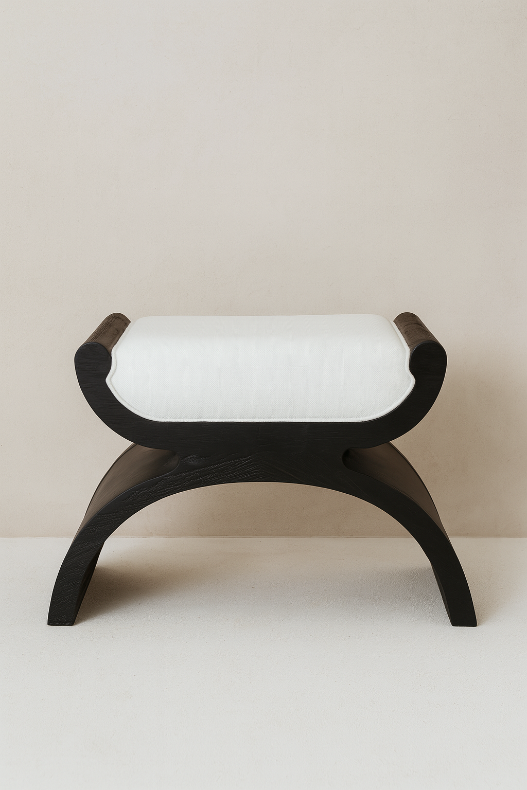 A sculptural wooden black ottoman with arched legs and a thick, upholstered cushion, set against a neutral-toned wall and concrete floor.