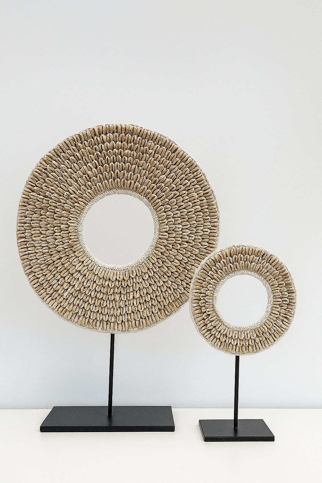 Two handmade cowrie shell discs on black metal stands against a light neutral wall, one large and one small