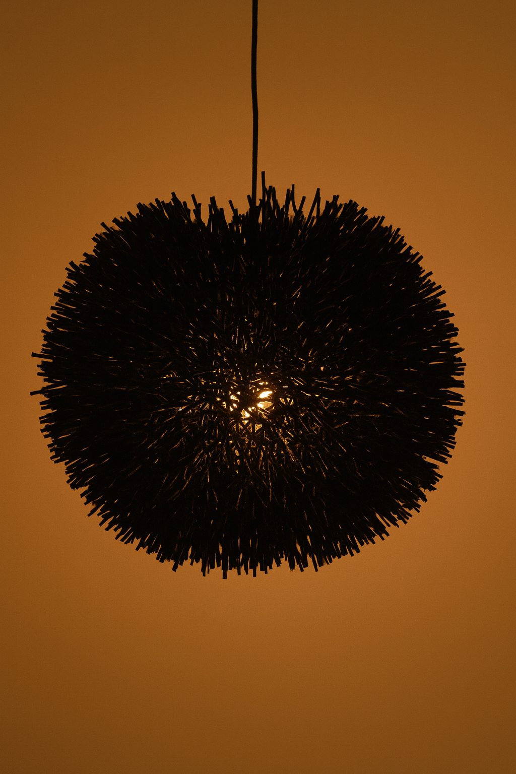 A black raffia pendant light in a spherical form, handwoven and glowing warmly against a neutral wall