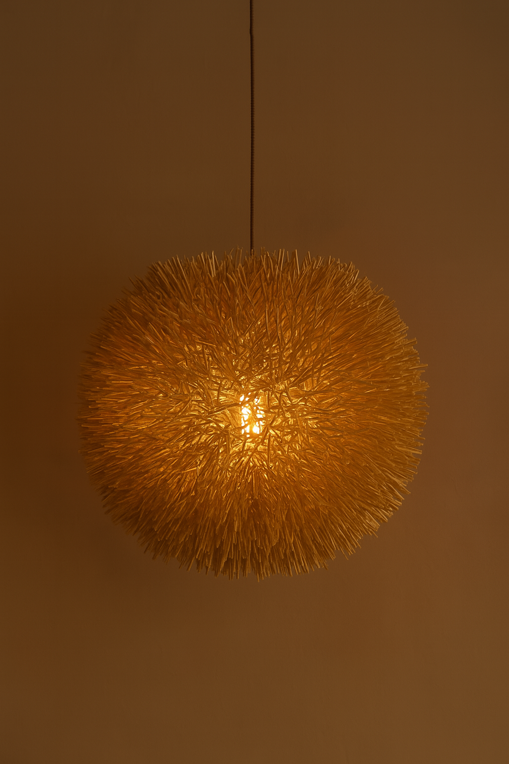 Handwoven spherical bamboo pendant light glowing softly against a warm brown wall – Jua Halo by Totem”