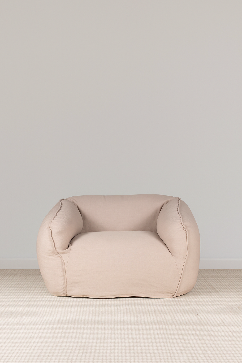 Soft rounded one-seater armchair in pale linen fabric, with deep cushions and curved edges, placed on a textured light rug against a white wall