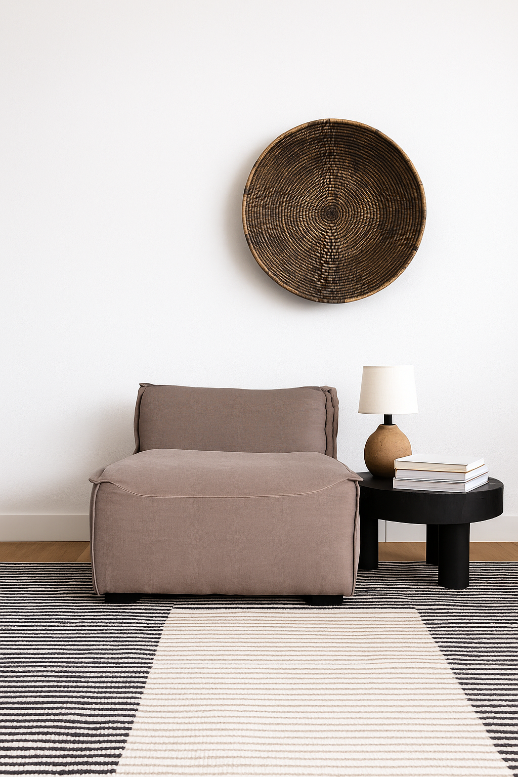 Low modern one-seater lounge chair with soft brown upholstery, styled on a striped rug with a black side table, ceramic lamp, and woven wall decor