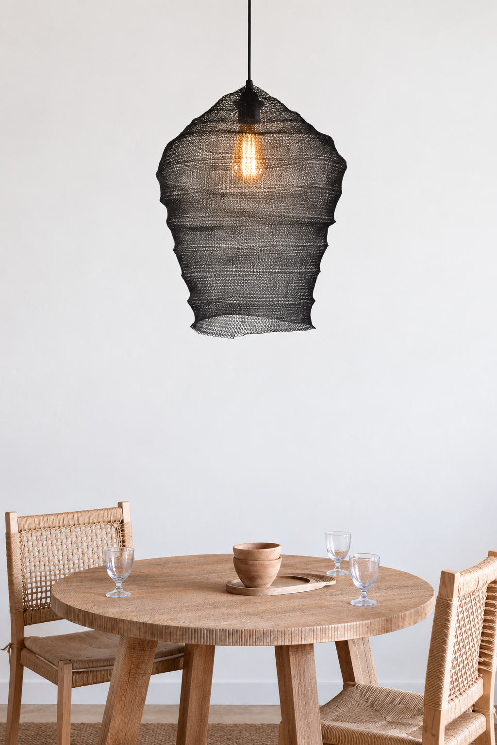 QORI Lampshade in black wire mesh with an organic tapered form and visible filament bulb, hanging over a round teak dining table with woven rattan chairs