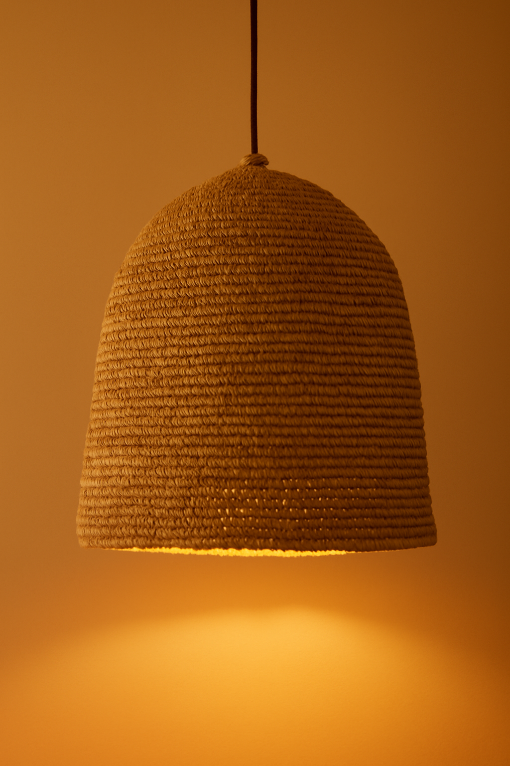 A photograph features a dome-shaped pendant lampshade handwoven from natural raffia, casting a warm ambient glow against a soft brown background.